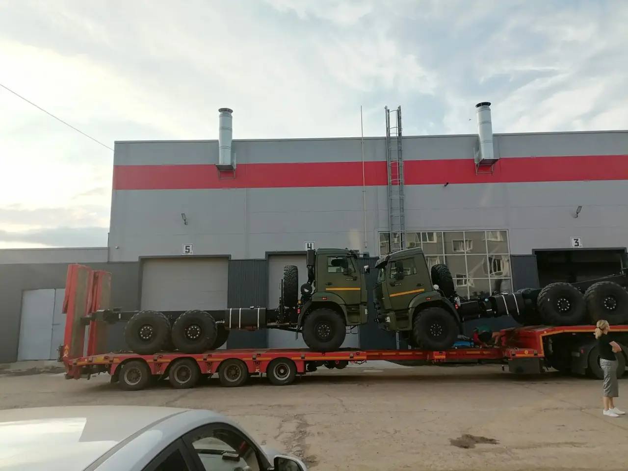 Два грузовика КАМАЗ цвета хаки закреплены кабинами друг к другу на красном низкорамном трале, вид сбоку у ворот производственного корпуса